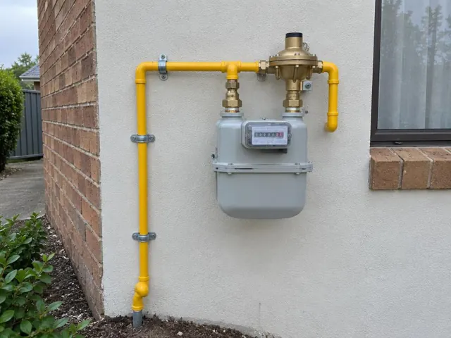 New yellow gas pipework and meter on an exterior rendered wall beside a brick window in Quakers Hill